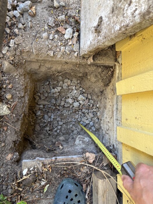 Grop utanför garage med grus och måttband som mäter djupet, intill gulmålad fasad och stenläggning, del av undersökning för elinstallation.