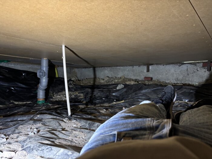 Person ligger i krypgrund med synlig elledning och rörsystem. Golvet täcks av svart plast och grus, med träskiva ovanför.
