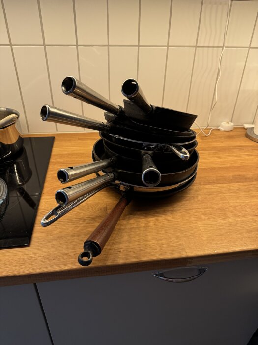 En stapel med olika stekpannor med handtag på en köksbänk, mot en kaklad bakgrund.