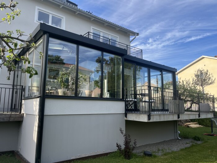 Glasinklädd terrass med svart metallräcke mot en vit putsad husvägg, omgiven av grönska och blå himmel.