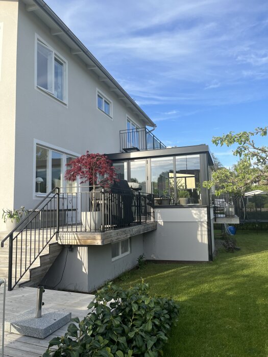 Vitputsat hus med altan på baksidan, omgiven av grönska och rött träd i kruka. Svart räcke och glasad verandadel samt kvällssol över trädgården.