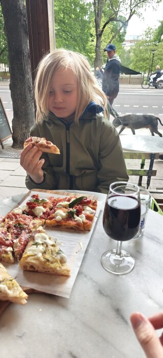 En person äter pizza vid ett bord utomhus, med fler pizzabitar på en skärbräda och ett glas rödvin bredvid.