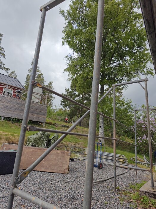 Byggställning under uppbyggnad med träram och metallstänger, omgiven av träd och en liten röd stuga i bakgrunden.