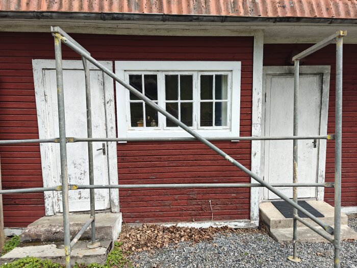 Röd byggnad med vit dörr och fönster, byggnadsställning framför.