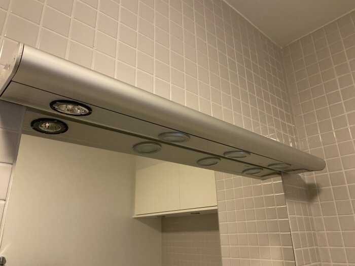 Belysningsramp med fyra spottar ovanför en badrumsspegel, monterade på väggen med synliga glödlampor och armaturer mot kakelplattor.