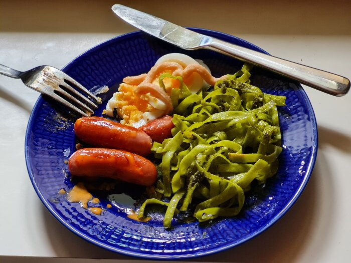 Prinskorv med sriracha mayo, kokt ägg med Kalles kaviar och spenattagliatelle med pesto på en blå tallrik.