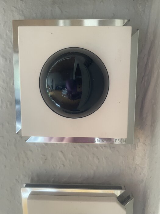 Bang & Olufsen-väggpanel med rund svart knapp och reflekterande yta, kan vara del av hemelektroniksystem i huset.