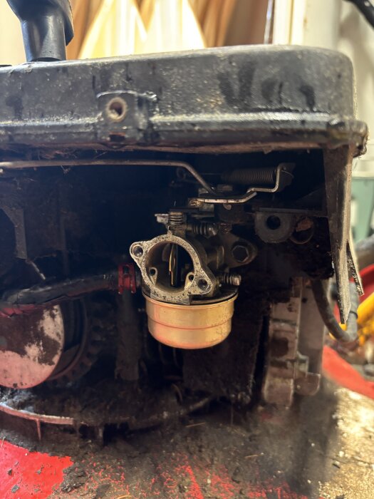 Närbild av en gräsklipparmotor med borttagen förgasare, synlig bränslekopp och reglage. Motorn är delvis smutsig och saknar luftfilter.