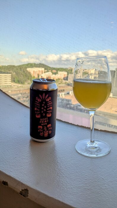 En burk Bootprint-öl från Hop Notch och ett vinglas med öl, ståendes på en fönsterbräda med utsikt över en stad.