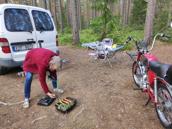 En person grillar grönsaker och korv vid en vit skåpbil i skogen, med campingutrustning och en röd moped i närheten.