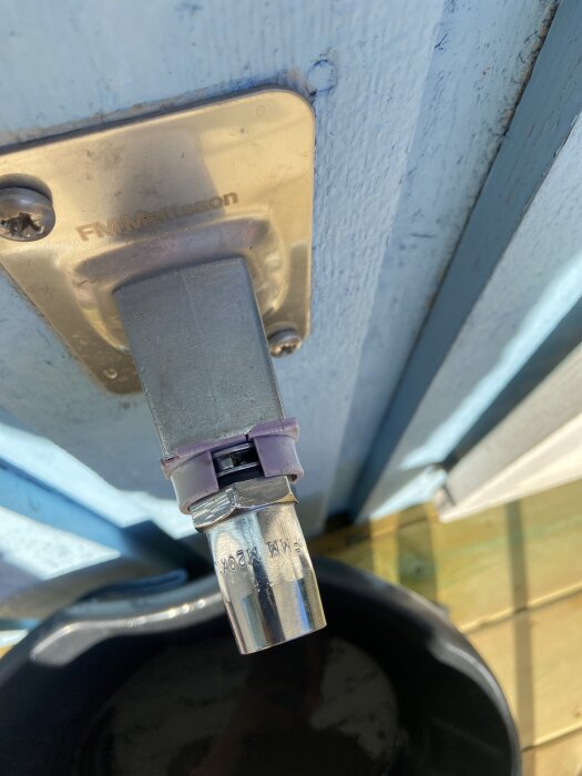 Utomhusvattenkran med läckage från ett litet hål nära anslutningen, monterad på en blå trävägg ovanför en svart hink.