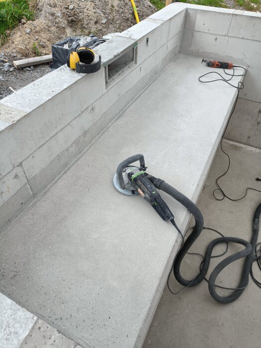 Betongbänk under slipning med en Festool-slipmaskin. Hörselkåpor och verktyg syns ovanpå bänken. Spricka synlig mellan bänk och vägg.