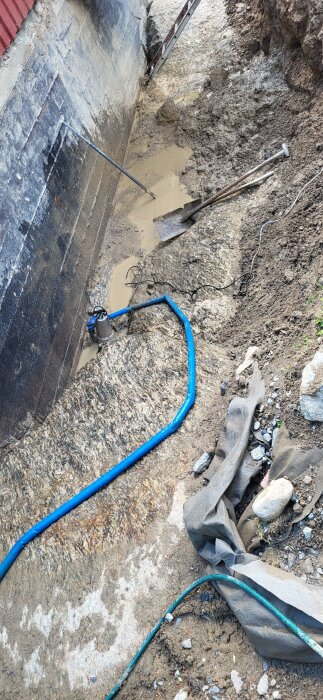 Bilden visar en dräneringsgrävning intill en husgrund på berg med verktyg som spade och slang samt lerigt vatten i botten av diket.