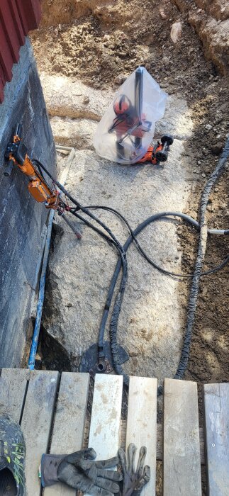 Dräneringsarbete intill ett hus på berg, med borrutrustning, kablar och verktyg. Arbetsredskap ligger på träplankor vid kanten av utgrävningen.