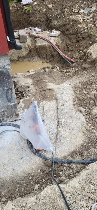 Gropar vid husgrund för dränering genom berg, med kablar i skyddsplast och vattenpölar, del av byggprojekt.