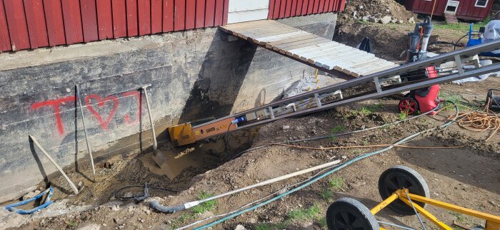 Dräneringsarbete vid ett hus på berggrund med grävd grop, transportband och verktyg; hjärta med bokstäver målade på husgrund.