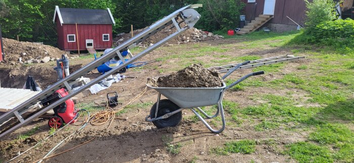 Skottkärra med jord framför transportband på en byggarbetsplats med en liten röd stuga i bakgrunden.