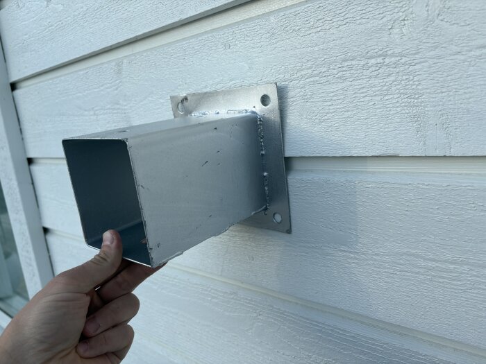 Montering av stolpfäste på husfasad inför bygget av pergola. En hand håller ett metallfäste mot en vit träfasad.