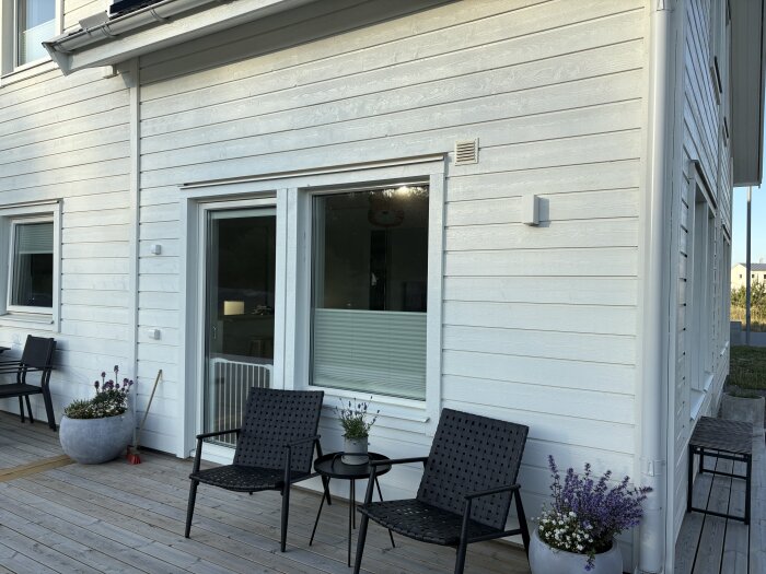 Vit husfasad med altandäck, två svarta stolar och blommor i krukor. Potentiellt område för pergola montering.