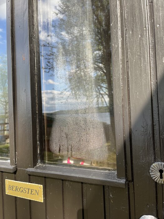 Ytterdörr med svart panel och två små fönster med kondens mellan glasrutorna, namnskylt med Bergsten.