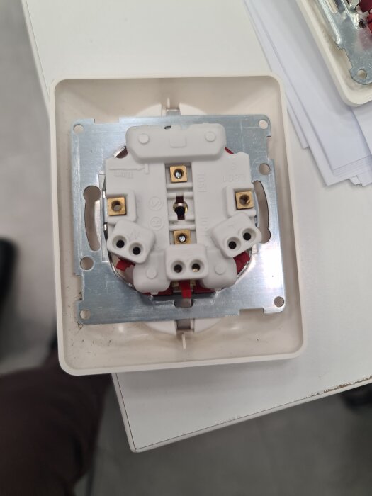 Närbild av ett elvägguttag utan kabelskruvar, med hål för blå och brun kabel samt jordning, monterat på en metallram.