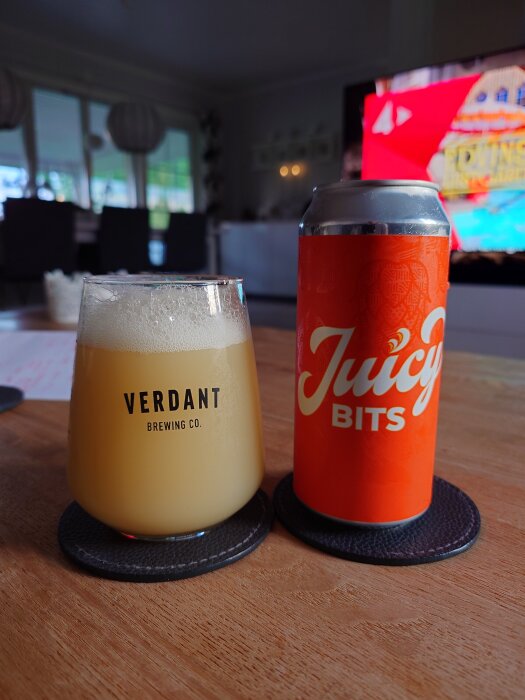 En burk öl med etikett "Juicy Bits" och ett glas med skummande öl från Verdant Brewing Co på ett bord.