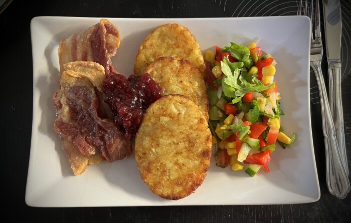 Stekfläsk med lingonsylt, tre potatisbullar och en färgglad sallad på en vit tallrik.