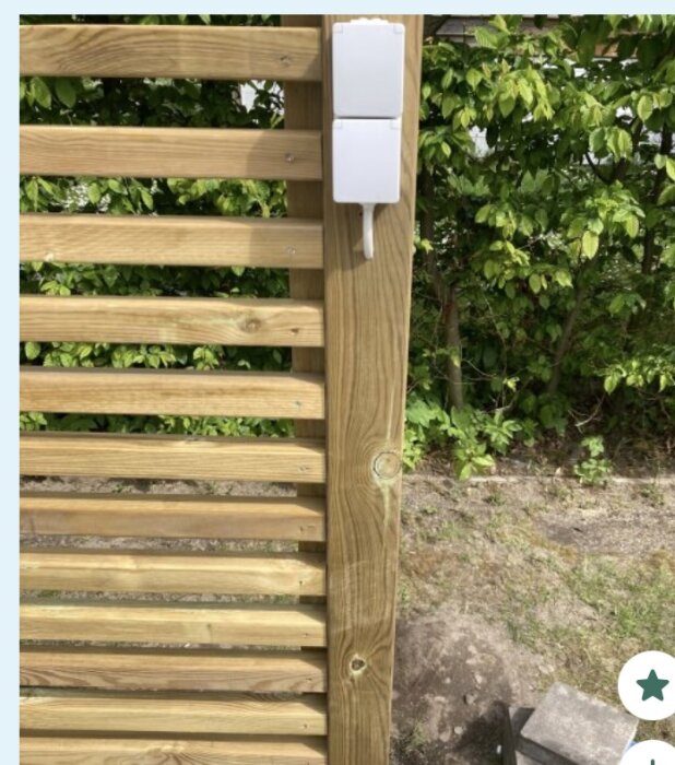 Uttag av typen Efapel IP65 monterat på ett trästaket, med sladd ansluten, i en trädgårdsmiljö.