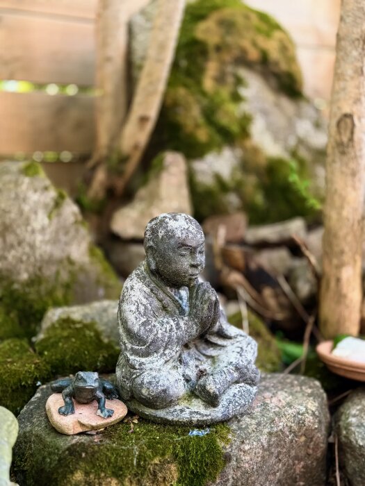 Staty av en mediterande figur i sten omgiven av mossa och en liten grodfigur på en sten, placerad i en trädgård.