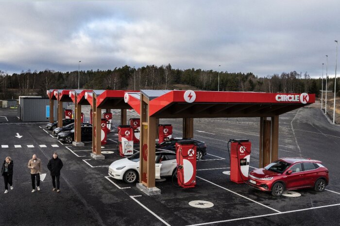 Elektrisk laddstation vid Circle K med flera parkerade bilar och personer som går mot laddningsområdet, omgiven av skog och öppen landskap.