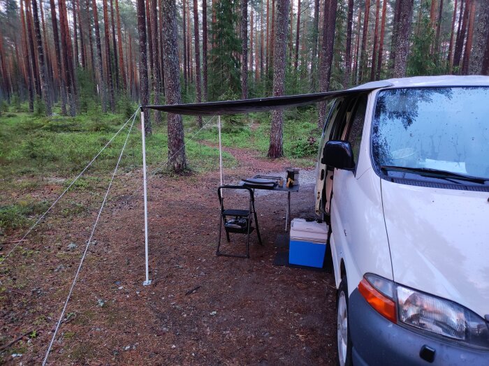 Bil med uppfälld markis i skog, bord och stol under, regndroppar synliga på fordonet, omgiven av träd.