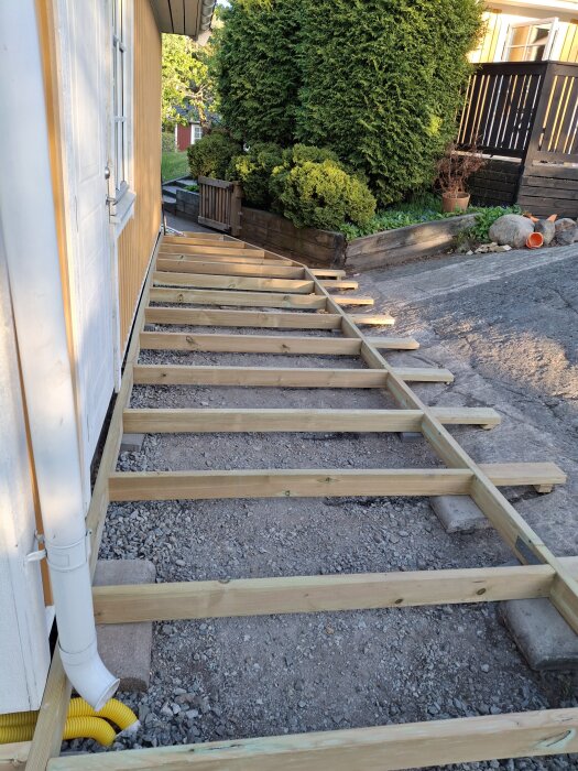 L-formad altan under uppbyggnad vid husvägg, stående på marksten och grus, med träreglar utan syllpapp mellan marksten och reglar.