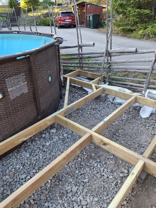 L-formad altanram av trä runt ett uppblåsbart poolområde, byggd på makadam och stenkross, utan grundisoleringspapp mellan reglar och marksten.