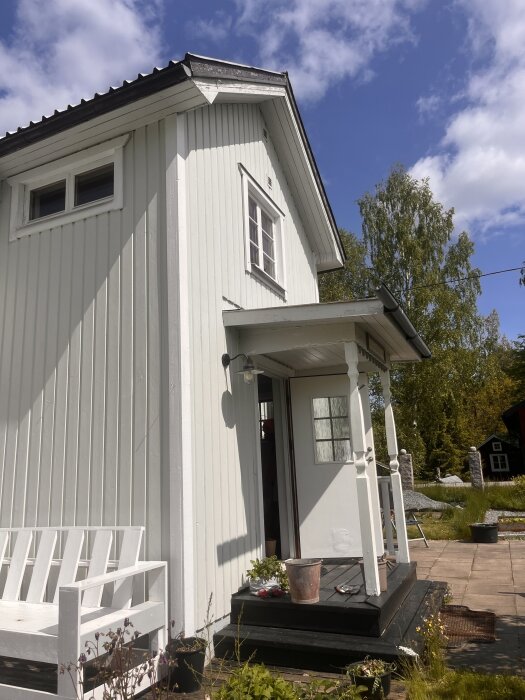 Vit trävilla med vindskivor i behov av målning, sett från en solig trädgård. Entrétak för lågt för att nå upp till vindskivorna.