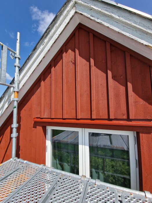 Röd träfasad med fönster och byggställning framför, under en klarblå himmel.