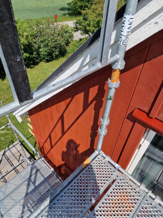 Byggställning vid röd träfasad, del av ett hus under renovering, med skuggor och tak i bakgrunden.