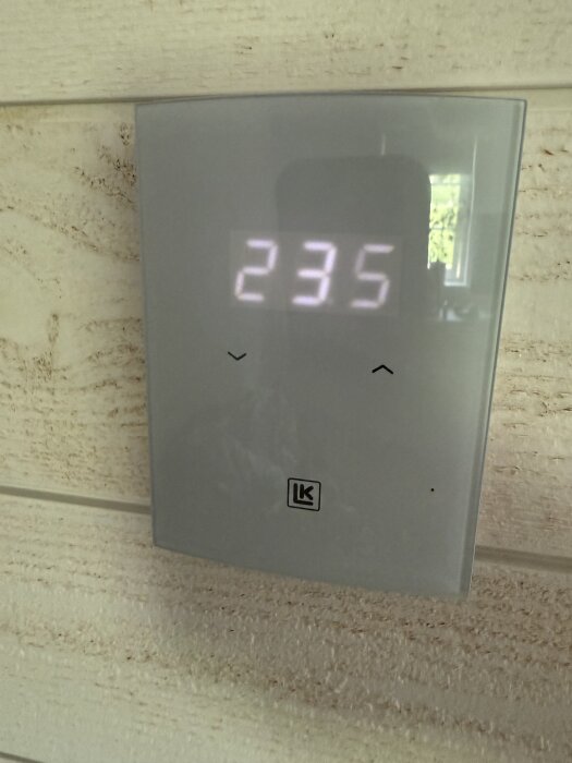 Digital termostat från LK med display som visar siffrorna 23,5. Monterad på en ljus trävägg.