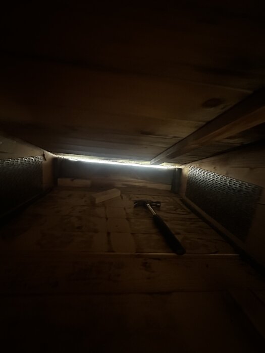 Utrymme på en vind med träbjälkar, delvis upplyst glipa mot taket, en hammare ligger på golvet.