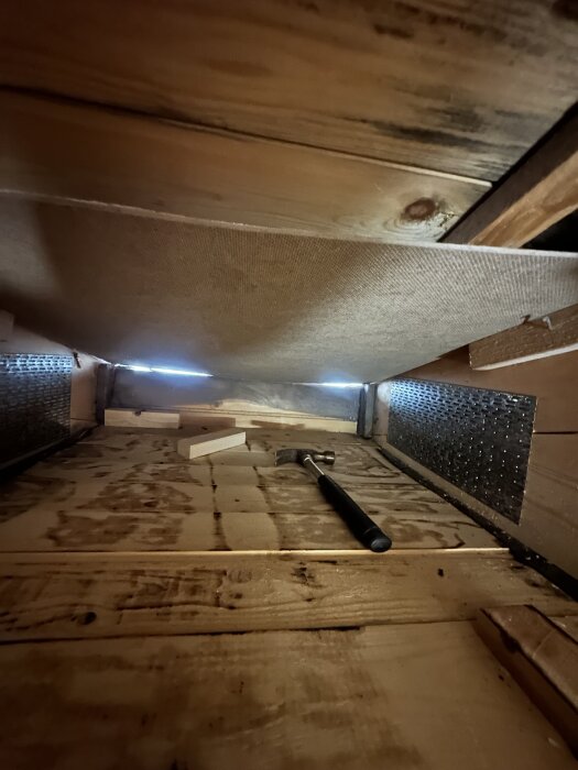 Utrymme på vinden med råspontgolv, träbitar, hammer och masonitskiva i taket, del av ventilationsarbete, synligt ljus vid takfoten.