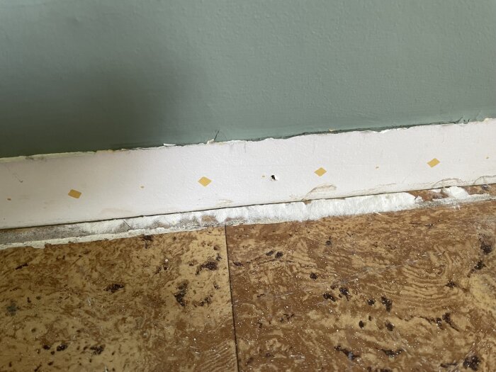 Vitt, pulveraktigt material syns under en borttagen tröskel i ett rum från 50-talet. Midgrön vägg och brunfläckigt golv i bakgrunden.