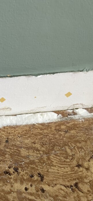 Vitt pulveraktigt material synligt under list i ett rum från 50-talet, med gröna och vita väggar samt trägolv.