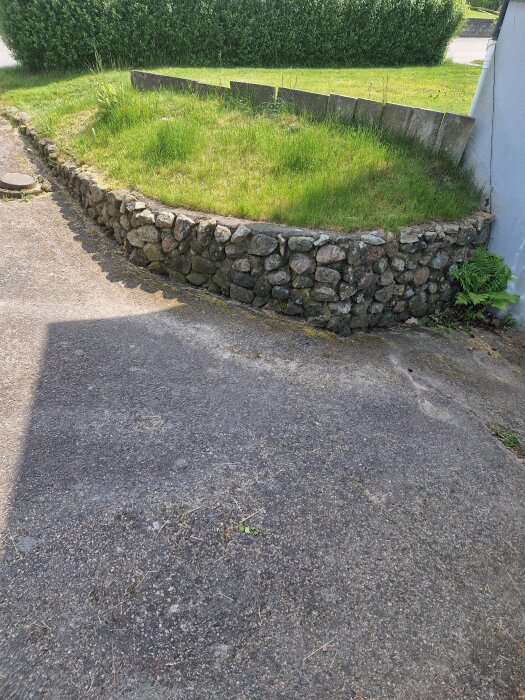 Stenmur vid gräsbevuxen sluttning, intill asfalterad yta och husvägg, med fokus på stödmurselement i ett trädgårdsprojekt.