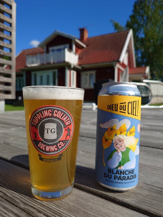 Glas och burk med veteöl på träbord framför ett rött hus i solsken.
