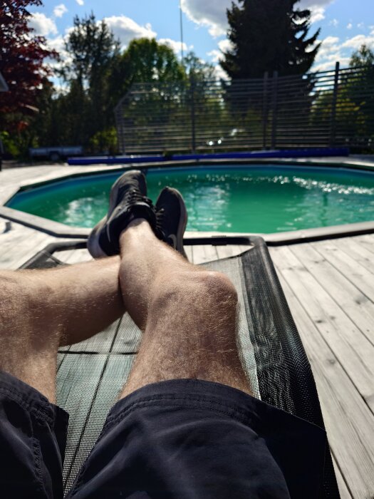 Person ligger på en solstol vid en rund pool med grönt vatten och blå himmel i bakgrunden.