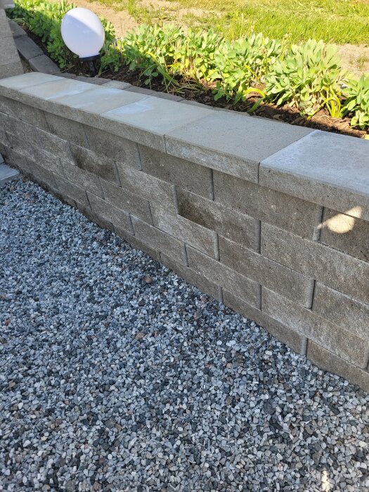 Stödmur med betongblock och geonät, planteringar ovanpå, grus framför och en rund solcellslampa.