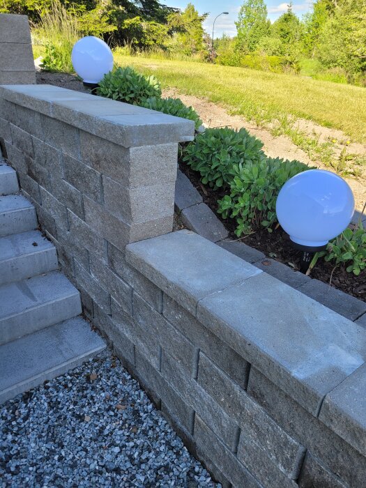 Murstenstrappa med kant och dekorationslampor i en trädgårdsanläggning, omgiven av grönska och stenar.