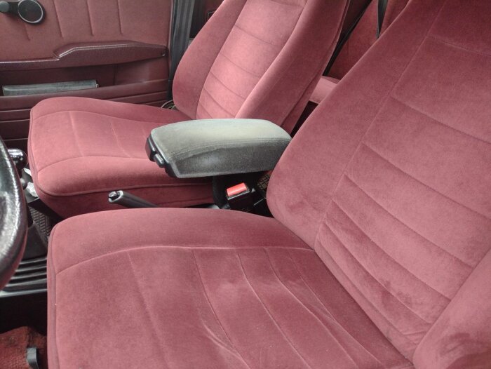 Röd bilinteriör med veloursäten och ett svartgrått armstöd av tyg mellan framsätena, med dryckeshållare.