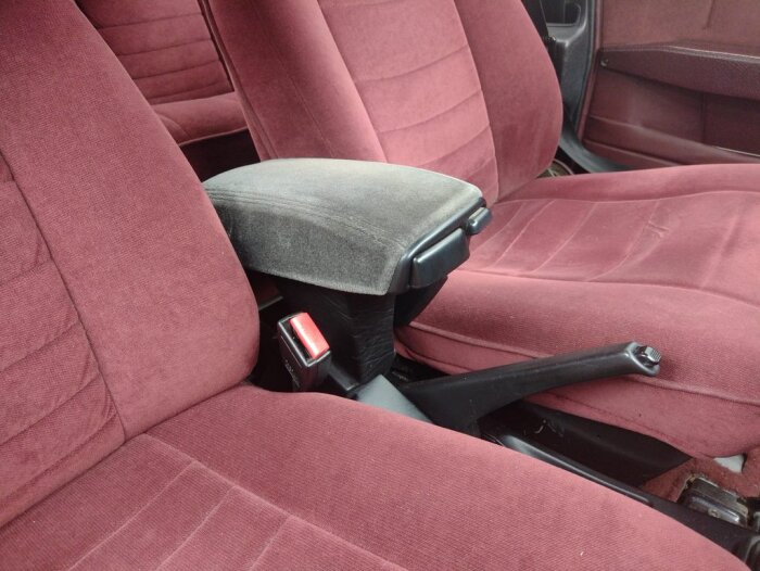 Armstöd i grått tyg installerat mellan två röda veloursäten i en bil, med en växelspak och säkerhetsbälte synliga.