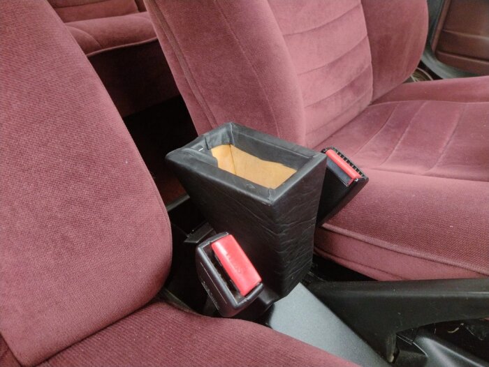 Hemmagjort armstöd i svart läder monterat i en röd Saab med velourklädsel, placerat mellan två bilsäten nära säkerhetsbältena.
