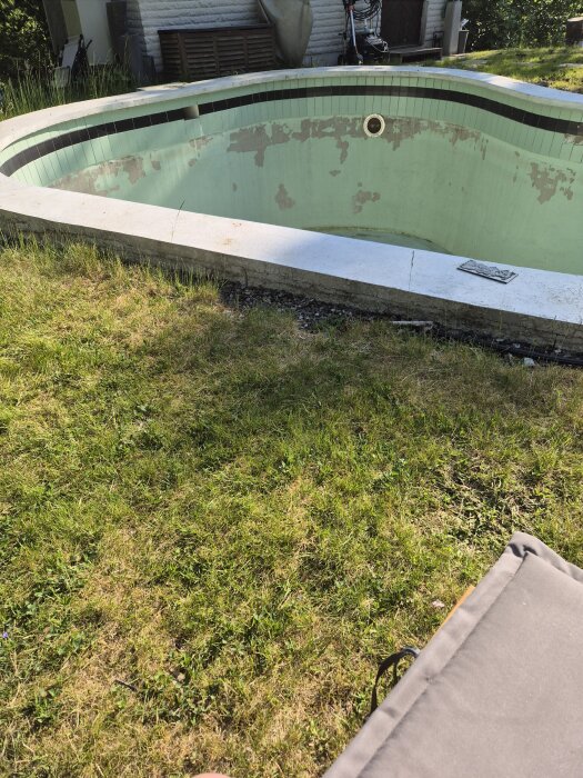 Gammal betongpool med flagnande färg och lösa kakelplattor. Poolen är torr och delvis omgiven av gräs och trädgårdsutrustning.